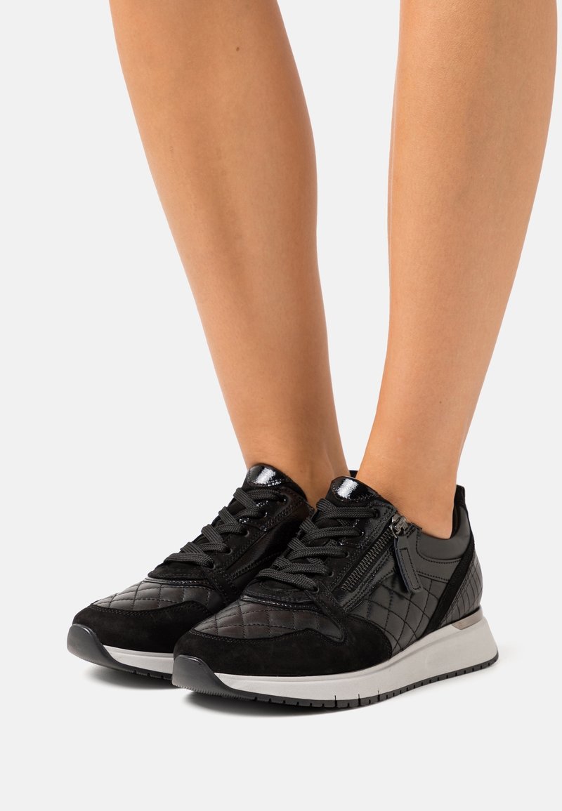 Jambes portant des baskets matelassées noires avec des lacets et des fermetures éclair latérales, debout sur une surface gris clair.