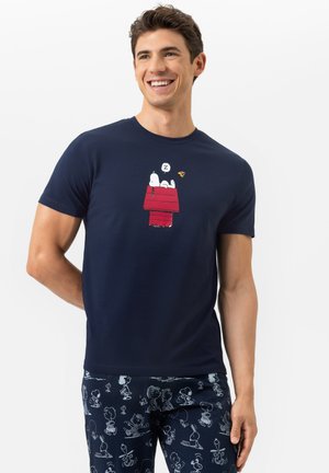 Ein lächelnder Mann trägt ein navyblaues T-Shirt mit Snoopy, der auf einem roten Hundehäuschen schläft, sowie dunkle Schlafanzughosen mit einem Muster aus Peanuts-Charakteren.
