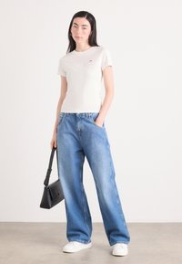 T-shirt bianco a maniche corte con piccolo logo, abbinato a jeans a gamba larga blu chiaro. La modella tiene una piccola borsa nera. Scarpe bianche.
