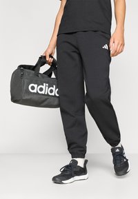 Μαύρο παντελόνι φόρμας Adidas με στενή εφαρμογή, που διαθέτει λευκό λογότυπο. Το μοντέλο κρατά μια γκρι τσάντα duffel της Adidas, σε συνδυασμό με μαύρα αθλητικά παπούτσια.