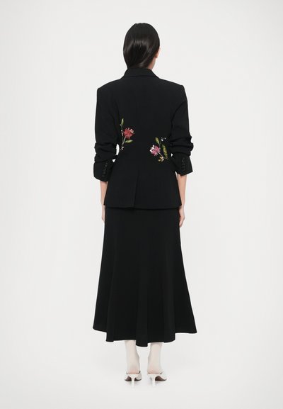 Fekete blézer piros és zöld hímzett virágdíszítéssel, egy légiesen omló fekete szoknyával párosítva. Fehér harisnya és magassarkú cipő fölött viselve.