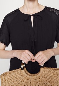 Blusa nera con accenti in pizzo, scollo a chiave e fiocco. Tiene in mano una borsa di paglia intrecciata con manico circolare e decorazioni di perline.