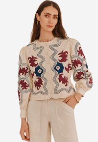 Blusa beige ricamata con vivaci design floreali in bordeaux e blu, scollo rotondo, chiusura con bottoni e maniche a sbuffo.