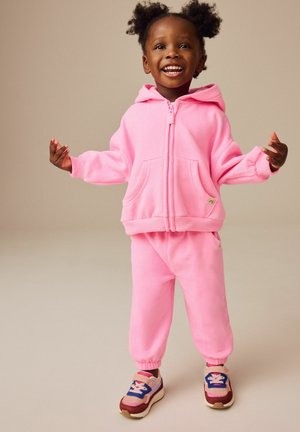 Roze hoodie en bijpassende joggers gemaakt van zachte, textuurachtige stof, voorzien van zakken en elastische boorden. Sneakers met meerkleurige accenten.