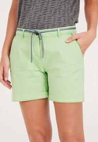 Lichtgroene shorts met opgerolde manchetten, voorzien van een trekkoord in de taille met blauwe accenten en zijzakken. Textuur grijs top zichtbaar erboven.
