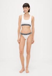Fehér bikini szett, amely crop topból és alacsony derekú alsóból áll. Fekete elasztikus pánt "justcavalli" logóval. Sima anyag, minimális textúrával.