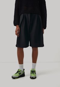 Shorts noirs mi-genoux en tissu lisse, associés à un pull ample en tricot bleu marine et des chaussures de sport gris-vert avec des accents néon.
