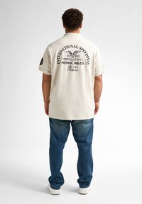 Beige poloshirt met achterop de print "INTERNATIONAL SPEEDWAY" en een adelaarsontwerp. Draagt met blauwe jeans en witte sneakers. Korte mouwen.