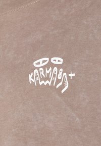 Light brown t-shirt with a textured fabric featuring a white graphic print of "KARMA" in a stylized, abstract font.