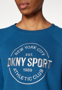 Blue sweatshirt with a round silver logo, featuring "DKNY SPORT," established 1989, and "ATHLETIC CLUB" in bold lettering.