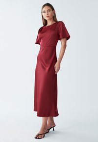 Bordo saten obleka s kratkimi rokavi, razširjenim robom, prilegajočim pasom in gladko teksturo. V kombinaciji z črnimi sandali na visoki peti z trakovi.
