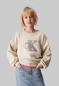 Sudadera beige con un gran logo plateado "cK" en la parte frontal. Combinada con una camiseta interior rosa y una falda de mezclilla azul, con un corte relajado.