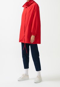 Rød vandtæt jakke med hætte, knaplukning, lange ærmer og justerbar snor i kanten, båret over mørke jeans og hvide sneakers.