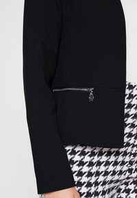 Veste noire à manches longues avec une poche zippée sur le côté. Le matériau semble lisse, avec un design structuré. Pantalon à motif pied-de-poule visible.