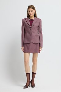 Jonge vrouw in een geprinte mauve blazer en bijpassende minirok, doorschijnende bordeaux rode sokken, spitse hakken, staand tegen een effen witte achtergrond.