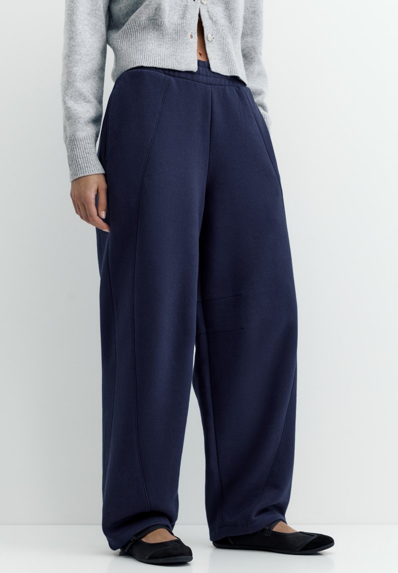 Person iført løstsiddende marineblå bukser, grå cropped cardigan og sorte flade sko, stående mod en helt hvid baggrund.