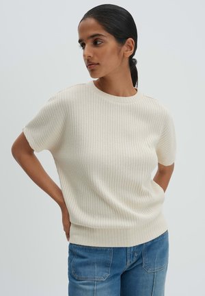 Donna con capelli scuri legati indietro che indossa un maglione bianco a maniche corte a coste e jeans di denim blu, posando con le mani sui fianchi.