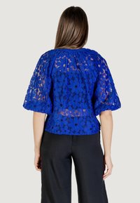 Top in pizzo blu con motivi floreali e maniche a sbuffo, dal taglio morbido e scollatura ampia. Abbinato a pantaloni neri per un contrasto.