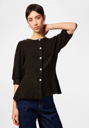 Jauna moteris su trumpais tamsiais plaukais dėvi juodą riboto stiliaus megztinį su sagomis ir tamsiai mėlynas džinsus, pozuodama su viena ranka pakelta už kaklo.