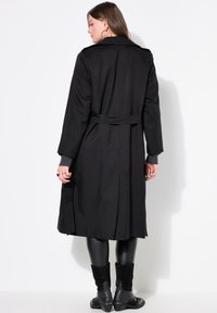 Manteau trench noir avec une taille ceinturée, des manches longues et un col cranté ; fabriqué dans un tissu lisse avec des coutures visibles et des fentes sur les côtés.