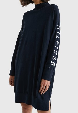 Kvinde iført en løs mørkeblå langærmet sweaterkjole med teksten ""TOMMY HILFIGER"" på ærmet og høj hals.