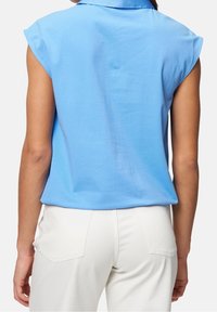 Chemise à manches courtes bleu clair avec un col, texture lisse et coupe ample, associée à un pantalon blanc avec des poches arrière visibles.