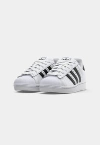 White Adidas Superstar sneakers with black stripes, rubber shell toe, and laces, shown side by side on a white background.
