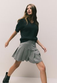 Czarny sweter dzianinowy z krótkimi rękawami, połączony z szaro-białą spódnicą w zygzak. Modelka stoi w aktywnej pozie.