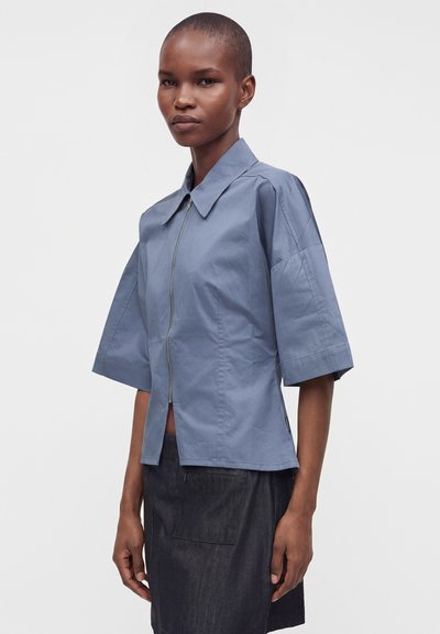Femme aux cheveux courts portant une chemise bleue à fermeture éclair avec des manches larges et une jupe foncée, debout devant un fond clair uni.