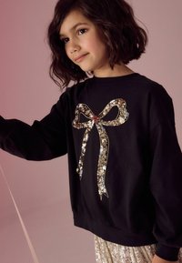 Svart sweatshirt med en stor, paljettdekorerad båge. Långa ärmar med rynkade manschetter, bärs över en glänsande, paljettklädd kjol.