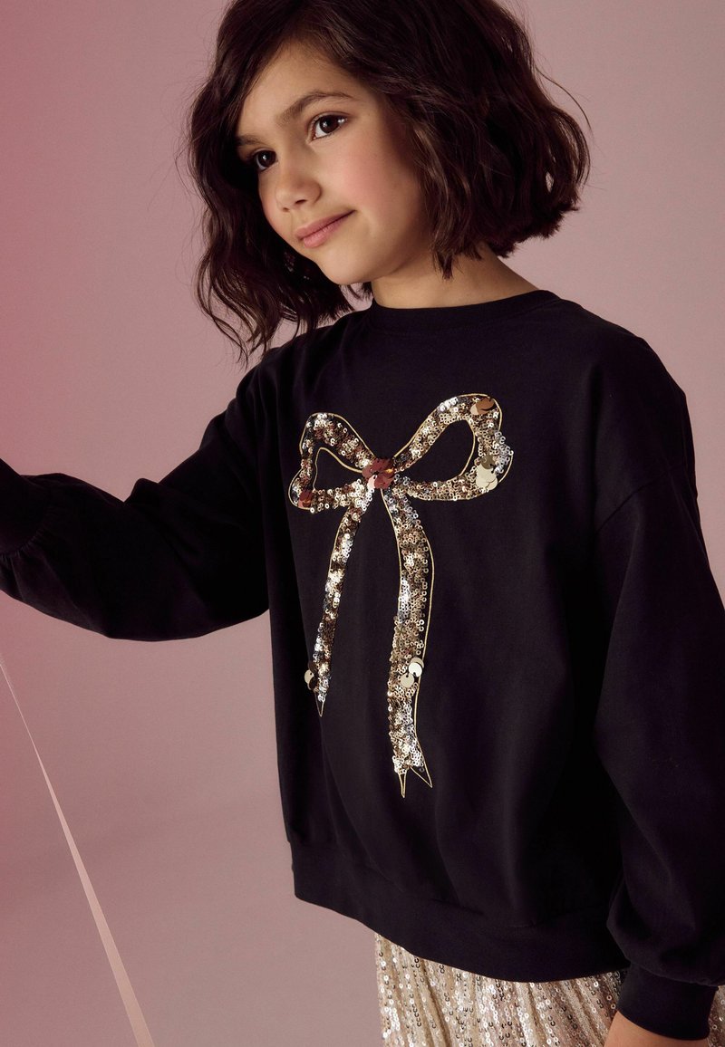 Svart sweatshirt med en stor, paljettdekorerad båge. Långa ärmar med rynkade manschetter, bärs över en glänsande, paljettklädd kjol.