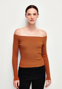 Suéter acanalado de hombros descubiertos en color óxido, con mangas largas, un diseño ajustado y una textura suave. Combinado con jeans oscuros.