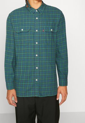 Homme portant une chemise à manches longues à carreaux verts et bleus avec poches poitrine avant et un pantalon noir, debout devant un fond uni.