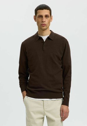 Jeune homme aux cheveux courts et foncés, portant un polo marron foncé à manches longues et un pantalon beige clair, debout avec une main dans la poche.