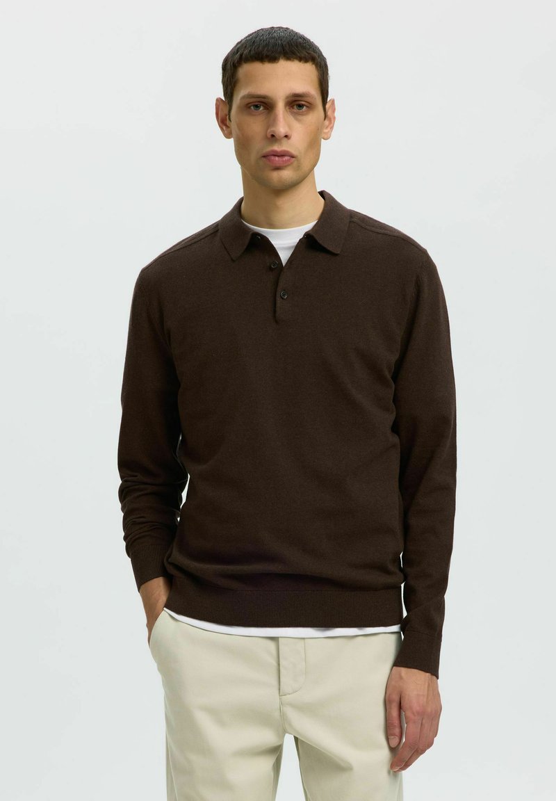 Jeune homme aux cheveux courts et foncés, portant un polo marron foncé à manches longues et un pantalon beige clair, debout avec une main dans la poche.