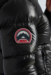 Lesklá čierna puffa bunda s rukávom, ktorý má okrúhlu nášivku s horským logom a nápisom "JACK1T" červeným písmom.