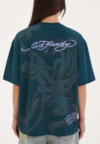 Teal oversized t-shirt med et stort, falmet sort grafisk design på ryggen og hvid "Ed Hardy" tekst i kursiv. Stoffet ser blødt ud.