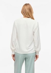 Femme aux longs cheveux blonds portant un chemisier blanc texturé à manches longues avec des poignets smockés et un pantalon large vert clair, vue de dos.