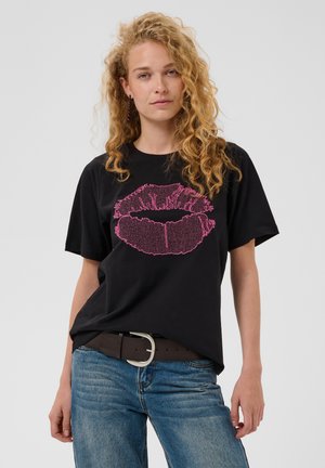 Jeune femme aux cheveux blonds bouclés portant un t-shirt noir avec une grande impression rose de lèvres et un jean bleu sur un fond uni.