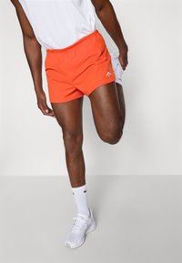 Orangene Sportshorts mit einem elastischen Bund und einem kleinen weißen Logo auf der Vorderseite; getragen mit weißen Sneakern und Socken.