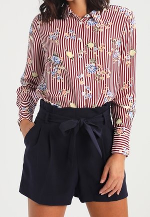 Mujer que lleva una camisa de flores a rayas rojas y blancas abotonada, metida en unos shorts azul marino de talle alto con cinturón de tela anudado.