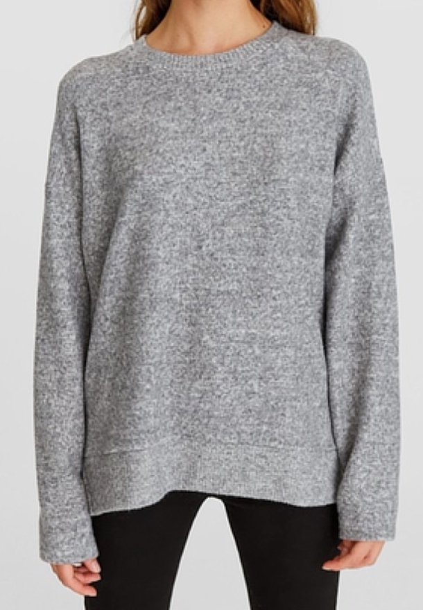 Femme portant un pull en maille gris clair à manches longues et un pantalon noir, debout devant un fond clair uni.