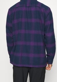 Violetti ja tummansininen ruudullinen pitkähihainen paita, jossa on napillinen suljenta ja pyöristetty helma, valmistettu pehmeästä flanellikankaasta.