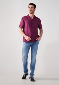 Rövid ujjú bordó ing, gombos, világoskék farmernadrággal és fekete sportcipővel párosítva. Az ingnek van gallérja és sima szövet textúrája.