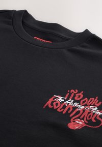 Sort bomulds-T-shirt med rund hals. Har et rødt og hvidt grafisk tekstdesign med teksten "it's only Rock'n Roll" og en tunge-logo.