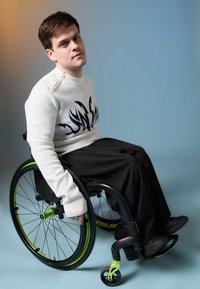 Λευκό πουλόβερ με σχέδιο μπλε φλόγας, μαύρο παντελόνι σε χαλαρή γραμμή και μαύρα παπούτσια. Η αναπηρική καρέκλα διαθέτει μαύρο σκελετό και πράσινες λεπτομέρειες.