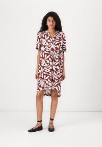 Robe florale rouge et blanche avec des manches courtes, devant à boutons, et une longueur d'ourlet arrière plus longue. Associée à des chaussures plates noires à brides.