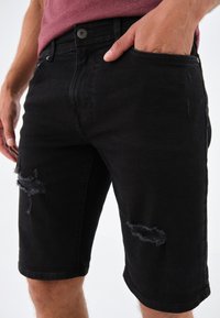 Shorts de mezclilla negra con un diseño ajustado, que presentan detalles desgastados y parches deshilachados en los muslos. Estilo clásico de cinco bolsillos.