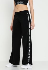 Pantalones de chándal anchos y negros con cintura ajustable, con el logotipo de "Reebok" en blanco a lo largo del costado. Fabricados con un material suave y transpirable.