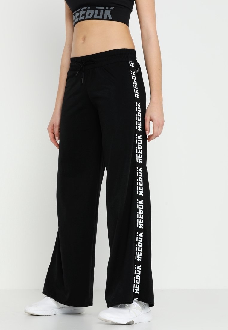 Pantalones de chándal anchos y negros con cintura ajustable, con el logotipo de "Reebok" en blanco a lo largo del costado. Fabricados con un material suave y transpirable.